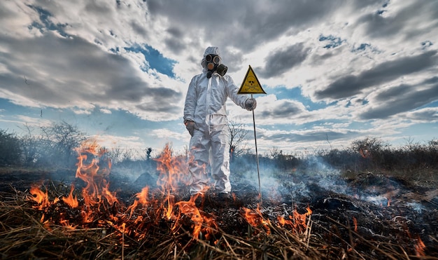 Feuerwehrmann Ökologe, der auf dem Feld Feuer bekämpft Mann in Schutzanzug und Gasmaske in der Nähe von brennendem Gras mit Rauch, der ein Warnschild mit Schädel und gekreuzten Knochen hält Naturkatastrophe-Konzept