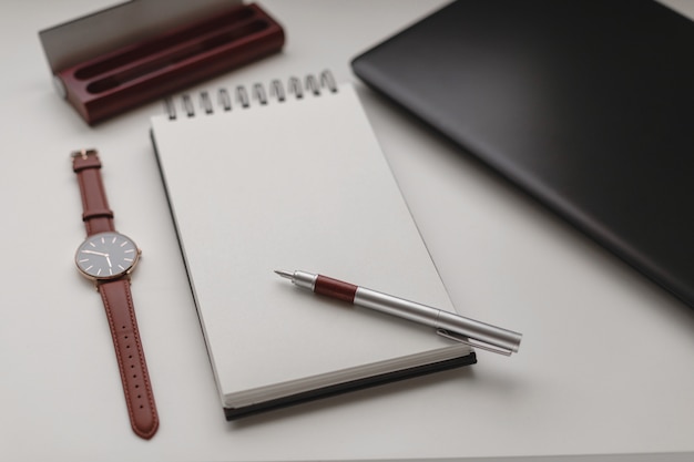 Öffnen Sie das weiße Spiralblock mit einem leeren Blatt und viel Bürozubehör, einer Armbanduhr, einem Mobiltelefon, einer Federtasche und einem Laptop.