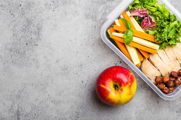 Fiambrera de alimentos saludables para oficina o escuela: filete de pollo, zanahoria, palitos de pepino, ensalada, manzana. Preparación y envasado de comida para apoyar un estilo de vida o dieta equilibrada, fondo de piedra, espacio de copia