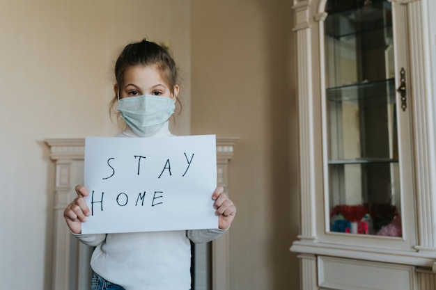 Ficar em casa conceito. Pequena menina na máscara segurando cartaz dizendo ficar em casa para proteção contra vírus e cuidar de sua saúde de COVID-19. Conceito de quarentena.