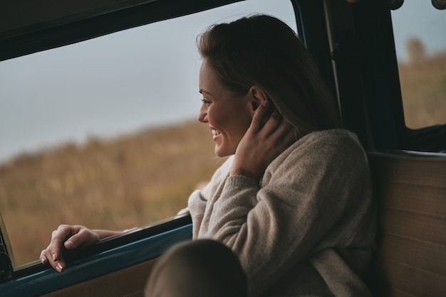 Fico feliz em viajar. Mulher jovem e atraente olhando pela janela da van e sorrindo enquanto está sentada no banco do passageiro da frente