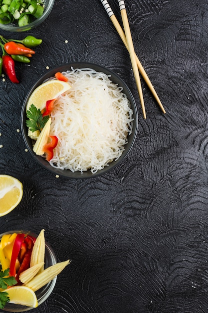 Fideos de arroz funchosa con verduras en un recipiente negro con palillos sobre un fondo oscuro, vista superior, flatlay.