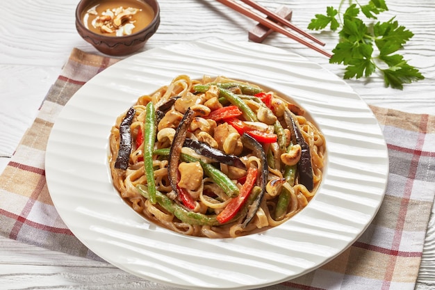 Fideos de arroz satay de pollo con judías verdes, pimienta y berenjena en un plato blanco sobre una mesa de madera, vista horizontal desde arriba, primer plano