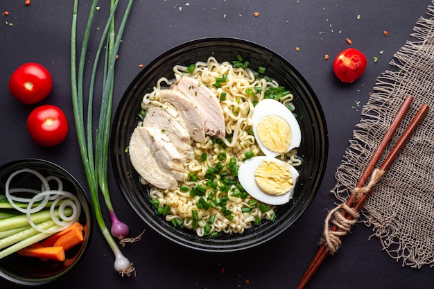 Fideos asiáticos de ramen con pollo, verduras y huevo en un tazón negro sobre fondo negro. Vista superior.