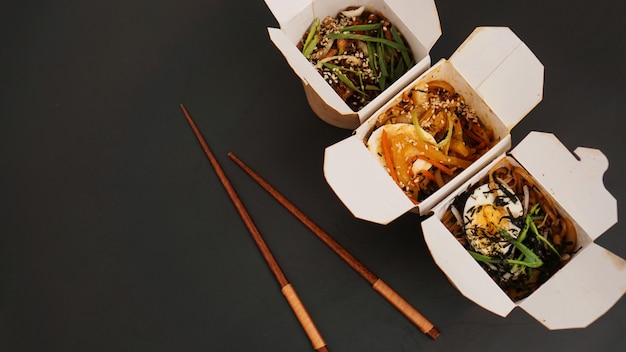 Fideos con carne de cerdo y verduras en caja de comida para llevar en el cuadro negro. Entrega de comida asiática. Alimentos en envases de papel sobre mesa negra