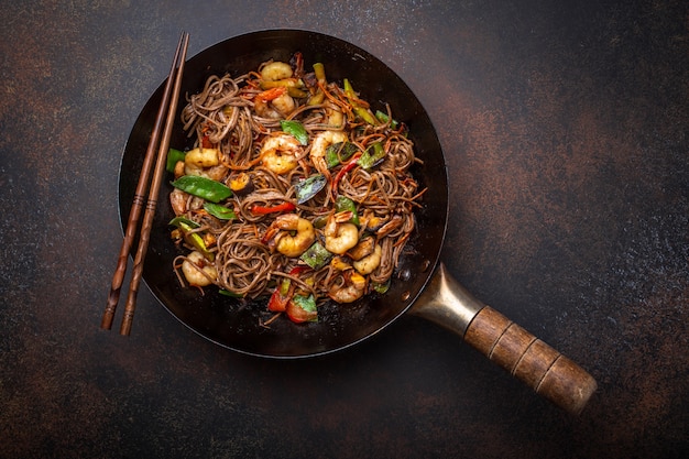 Fideos chinos soba salteados con camarones, verduras en una antigua sartén wok rústico servido sobre fondo de hormigón, de cerca, vista superior. Plato tradicional asiático / tailandés, cerrar