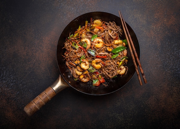 Fideos chinos soba salteados con camarones, verduras en una antigua sartén wok rústico servido sobre fondo de hormigón, de cerca, vista superior. Plato tradicional asiático / tailandés, cerrar