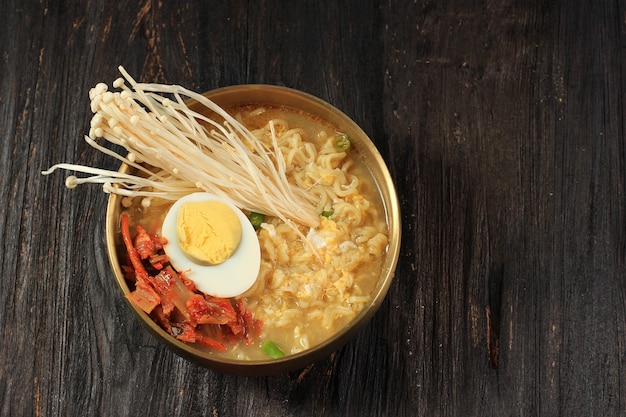 Fideos instantáneos estilo coreano, Ramyeon o Ramyun con sabor picante cubierto con medio huevo cocido, hongo Enoki y kimchi en un cuenco coreano dorado sobre una mesa de madera negra, aislado