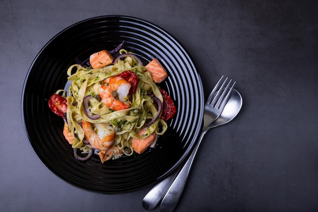 Fideos con mariscos, tomates secos, alcaparras y cebollas rojas Pasta casera con camarones, salmón, trucha y salsa pesto Fondo negro Placa negra Primer plano