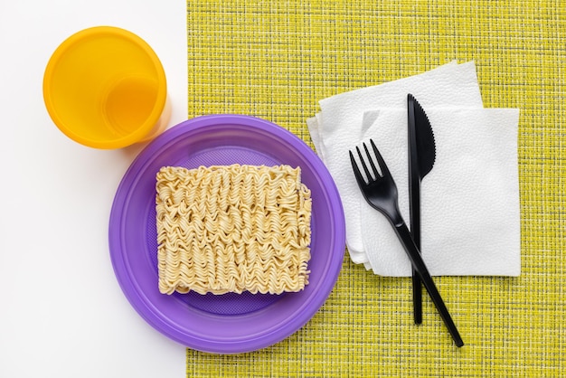 Fideos en un plato lila, vidrio plástico, tenedor y cuchillo negros, vista superior.