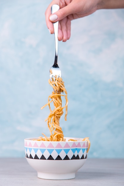 Fideos con salsa en un recipiente de color, la mano sostiene un tenedor de pasta colgando, espaguetis de apetito en azul.