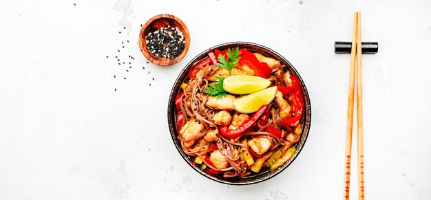 Fideos salteados de cocina asiática con pollo y verduras pimentón calabacín y semillas de sésamo en un tazón Espacio de vista superior de fondo de mesa de cocina blanca para texto