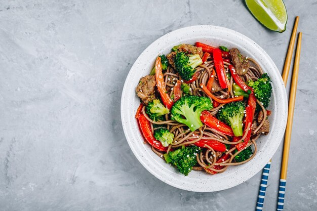 Fideos de ternera salteados con brócoli, zanahorias y pimientos rojos sobre fondo de piedra gris