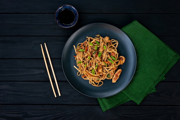 Fideos udon asiáticos con pollo con verduras y salsa teriyaki sobre un fondo negro de madera