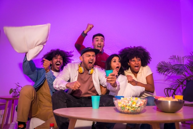 Fiesta de adultos Jóvenes sentados en el sofá viendo un partido de fútbol con palomitas Emoción por el fútbol led violeta celebrando un gol