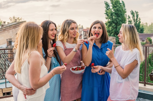 Fiesta de chicas. Hermosas mujeres amigas en el balcón divirtiéndose en la despedida de soltera. Ellos comen fresa