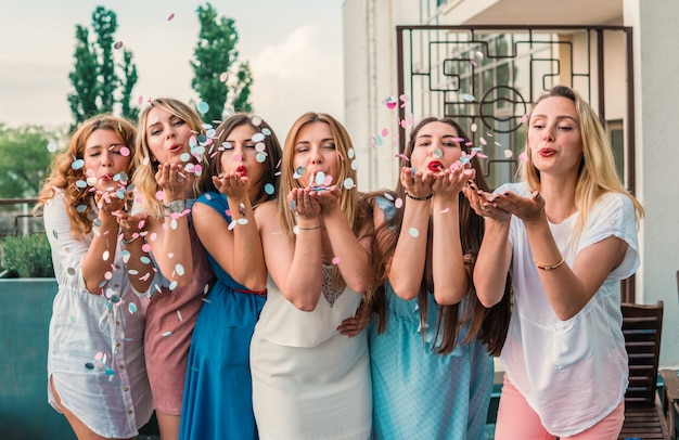 Fiesta de chicas. Hermosas mujeres amigas en el balcón divirtiéndose en la despedida de soltera. Están soplando confeti de las manos