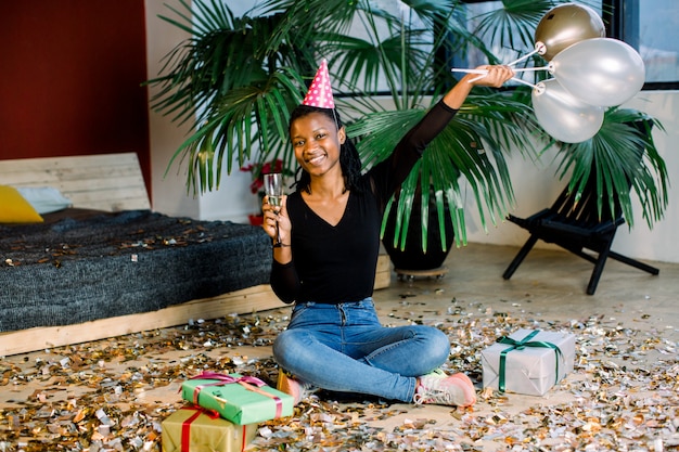 Fiesta de cumpleaños, carnaval de año nuevo. Mujer sonriente africana joven que celebra evento brillante ,. Confeti chispeante, divertirse, bailar y beber champán