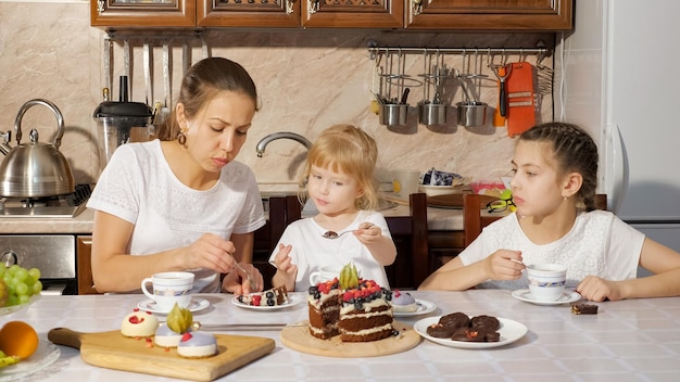 Fiesta de cumpleaños familiar en casa, mamá y dos hijas están tomando un té con pastel de chocolate casero en la cocina y riendo.