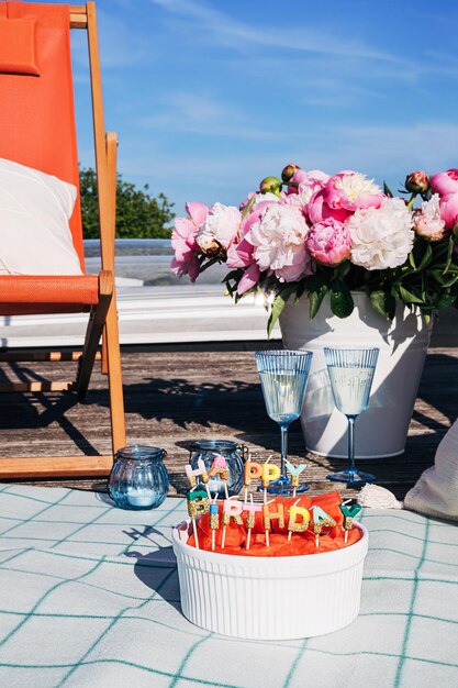 Fiesta de cumpleaños feliz de verano en el techo con manta de picnic fruta de sandía con velas copas de champán y sillón contra el cielo azul claro Idea de celebración al aire libre