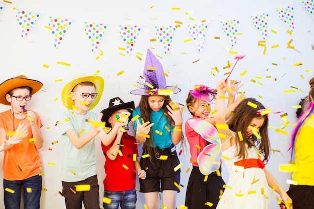 Fiesta de cumpleaños con niños en edad escolar con camisetas coloridas y sombreros elegantes y pelucas riéndose bajo confeti cayendo Niños soplando cuernos de fiesta celebrando el cumpleaños de su amigo