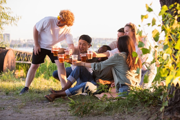 Fiesta grupo de amigos tintineo de vasos de cerveza durante un picnic en la playa bajo el sol