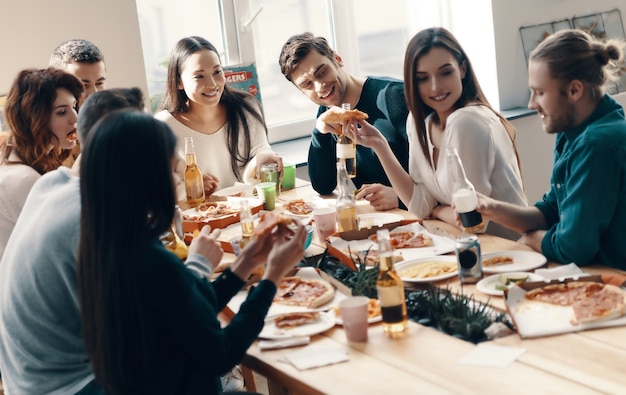 Fiesta inolvidable. Grupo de jóvenes en ropa casual comiendo pizza y sonriendo mientras tienen una cena en el interior