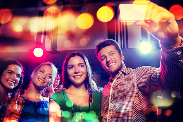 fiesta, vacaciones, tecnología, vida nocturna y concepto de personas - amigos sonrientes con copas de champán y smartphone tomando selfie en club nocturno con luces navideñas