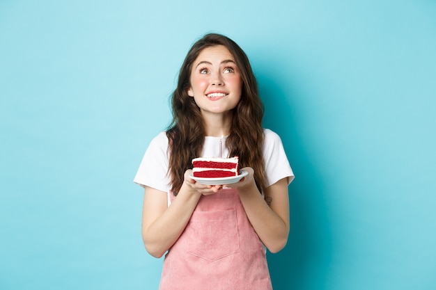 Fiestas y celebraciones. Cumpleañera soñadora pidiendo deseos y mirando esperanzado, sosteniendo el pastel de cumpleaños y sonriendo, de pie sobre fondo azul.