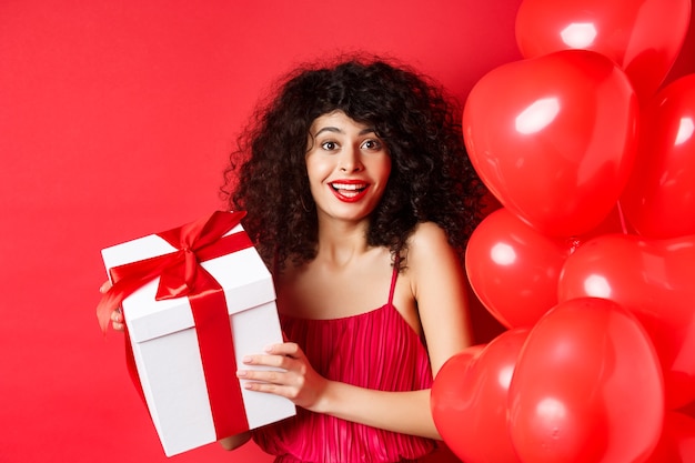 Fiestas y celebraciones. Hermosa mujer con cabello rizado, de pie cerca de globos de corazón, sosteniendo una caja de regalo y sonriendo feliz, fondo blanco.