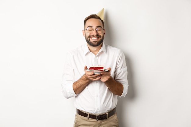 Fiestas y celebraciones. Hombre feliz con fiesta de cumpleaños, pidiendo deseos en b-day cake y sonriendo, de pie contra el fondo blanco.