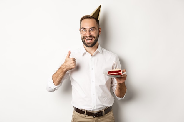 Fiestas y celebraciones. Hombre satisfecho disfrutando de la fiesta b-day, sosteniendo el pastel de cumpleaños y mostrando el pulgar hacia arriba en señal de aprobación, recomendando algo, fondo blanco.