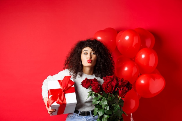 Fiestas y celebraciones. Mujer bonita celebrando cumpleaños soplando aire beso, recibir regalos y flores en aniversario, de pie cerca de globos de fiesta, fondo rojo