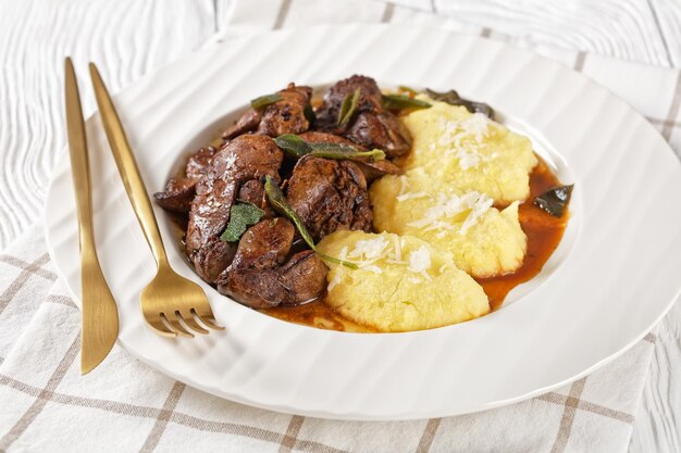 Fígados de frango com sálvia servidos com polenta em um prato branco na mesa de madeira clara Fegatini di pollo alla salvia cozinha italiana closeup