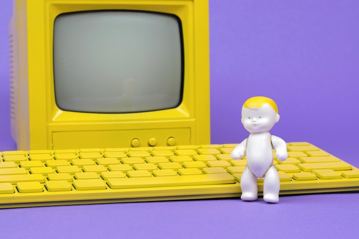 Foto una figura de bebé cerca de un teclado amarillo y un monitor amarillo sobre un fondo violeta.