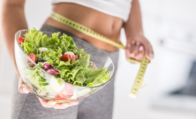 La figura deportiva de una atractiva mujer sosteniendo un tazón de ensalada de verduras frescas y cinta métrica.