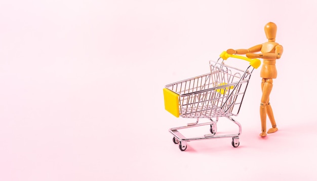 Figura humana de madera con un coche de compras en miniatura aislado sobre un fondo rosa