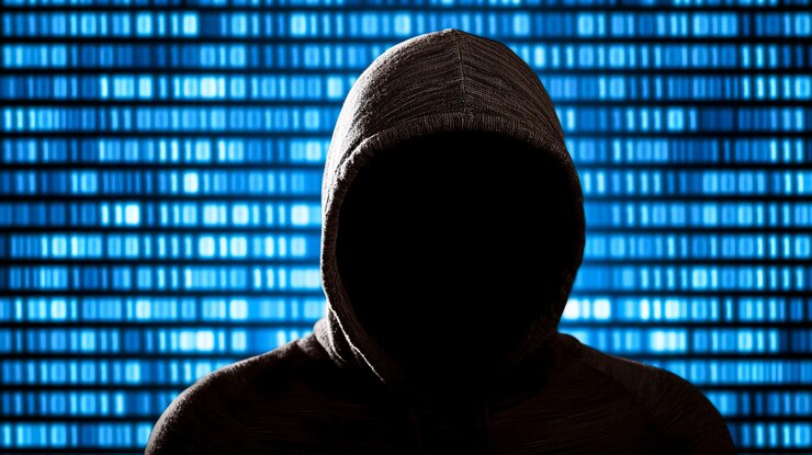 Foto figura misteriosa con sudadera con capucha oscura contra un fondo de código binario azul que representa amenazas digitales y desafíos de ciberseguridad en el mundo moderno