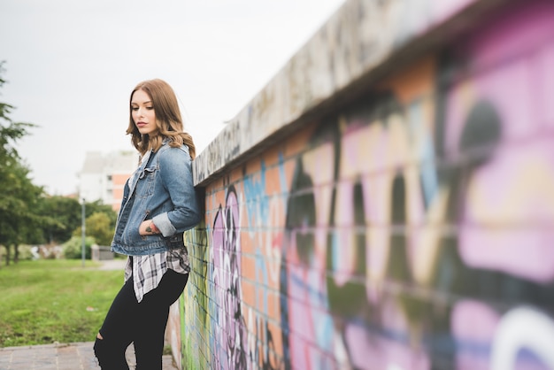 Figura de la rodilla de la mujer rubia caucásica hermosa joven del pelo recto que presenta inclinarse contra una pared