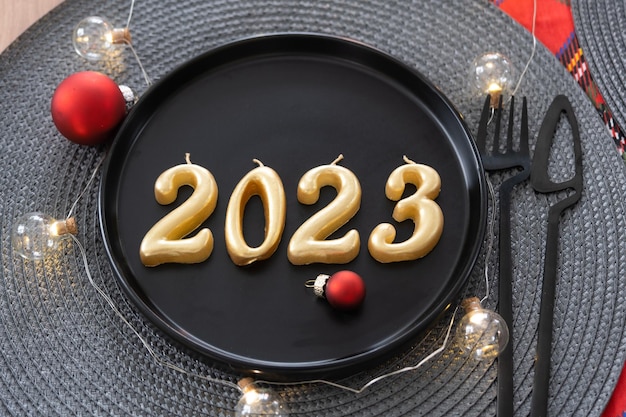 Figuras douradas 2023 de velas em um prato preto em uma mesa festiva com uma porção de Ano Novo Loftstyle festa interior festa Guardanapo de vime garfo faca