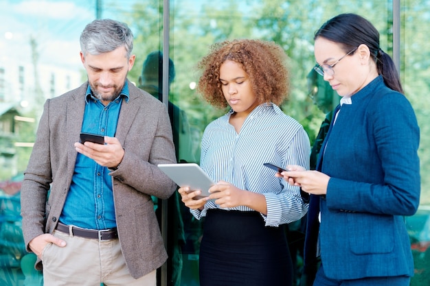 Fila de funcionários jovens e elegantes com dispositivos móveis, organizando o trabalho ou navegando na Internet ao ar livre