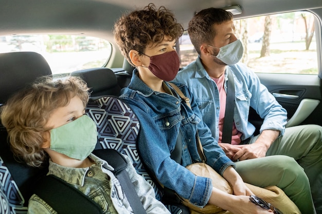 Fila de jovem família contemporânea de três pessoas com máscaras protetoras e jaquetas jeans olhando para a frente enquanto se dirige de carro no banco de trás