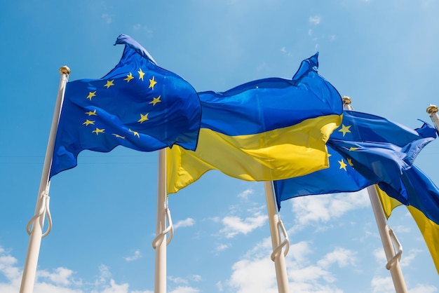 Fila de mastros de bandeira com bandeiras da União Europeia e Ucrânia tremulando pelo vento no fundo do céu azul