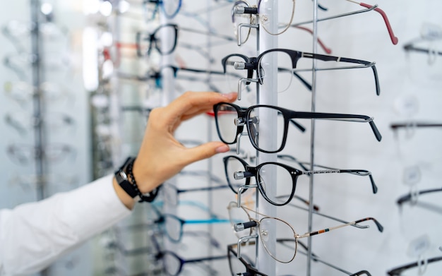 Fila de gafas en una óptica. Tienda de anteojos. Stand con gafas en la tienda de óptica. La mano de la mujer elige gafas. Corrección de la vista.