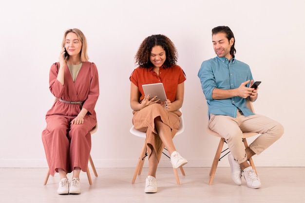 Foto fila de jóvenes amigos interculturales en ropa casual sentados a lo largo de la pared blanca y comunicándose a través de teléfonos inteligentes y touchpad