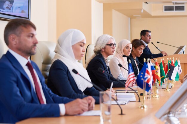 Fila de jóvenes delegados interculturales en ropa formal sentados a la mesa delante de micrófonos en la conferencia