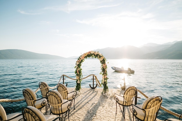 Fila de sillones delante de un arco de boda en un muelle adoquinado con el mar y las montañas en el