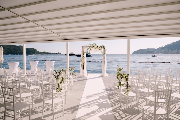 Filas de sillas blancas parados frente a un arco de boda bajo un dosel blanco a la orilla del mar