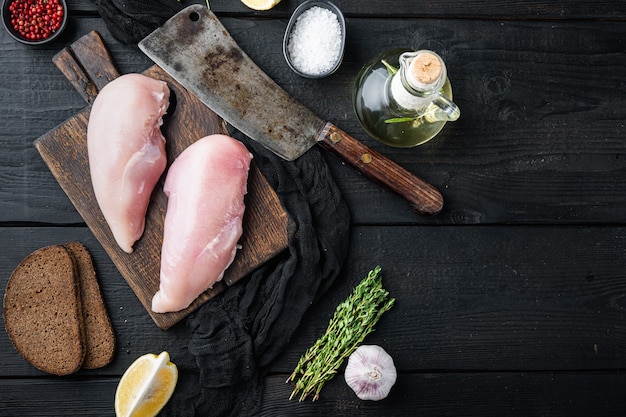 Filé de frango cru com ingredientes e faca de açougueiro na mesa de madeira preta, vista superior