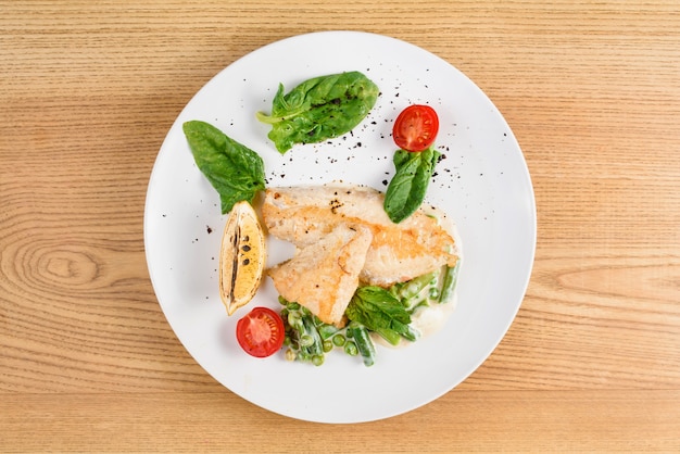 Filé de frango grelhado com manjericão, tomate cereja, limão, ervilhas, molho em prato redondo branco na mesa de madeira